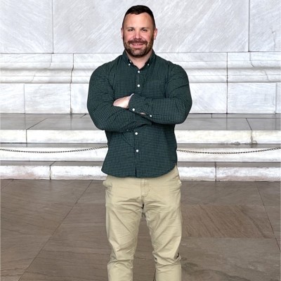 Dave Kaplan, a man with a short beard and muscular build, wearing a green gingham shirt and khaki pants, standing with arms crossed in front of a stone building.